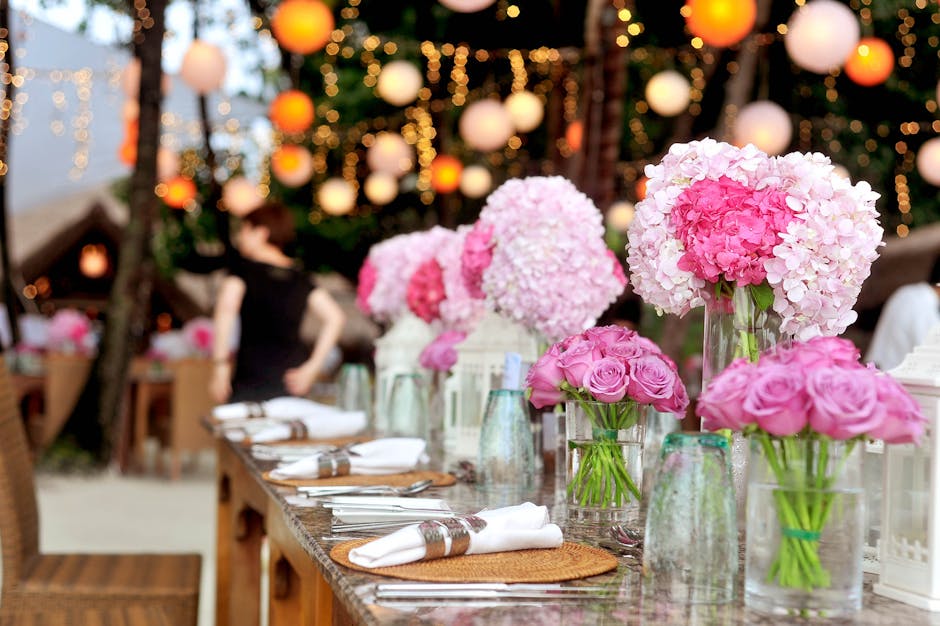 A long wooden table set for a floral wedding reception, decorated with large, lush bouquets of pink and white hydrangeas and roses in clear glass vases. The table is arranged with neatly folded white 