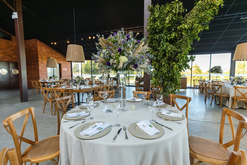 A round table in a wedding reception venue is decorated with a tall, elegant floral arrangement featuring white, purple, and lavender flowers, including roses and lavender sprigs, set in a clear glass