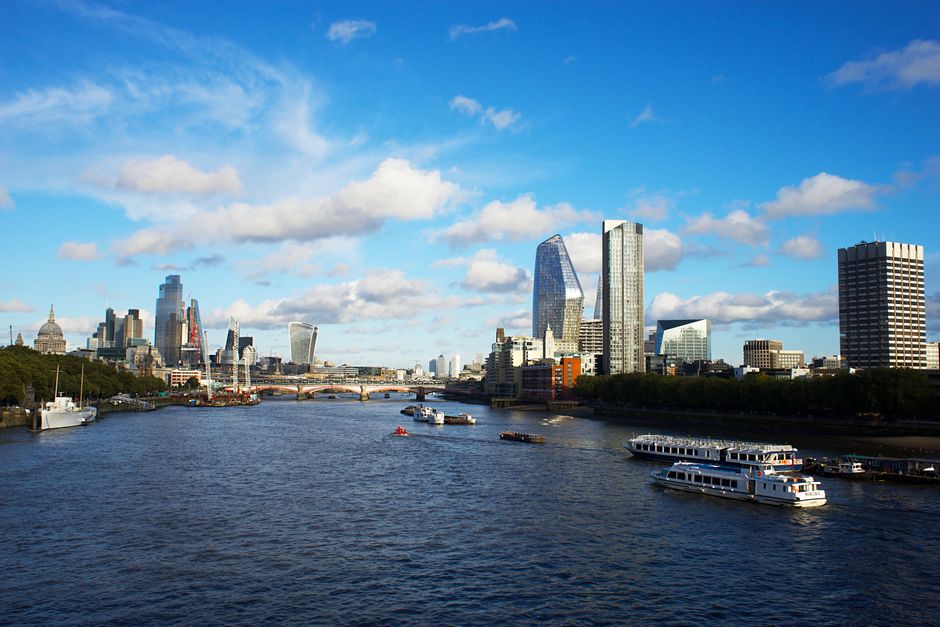 A scenic view of the River Thames in London with several modern skyscrapers in the background, including the distinctive Gherkin and Walkie Talkie building, under a bright blue sky with scattered fluf