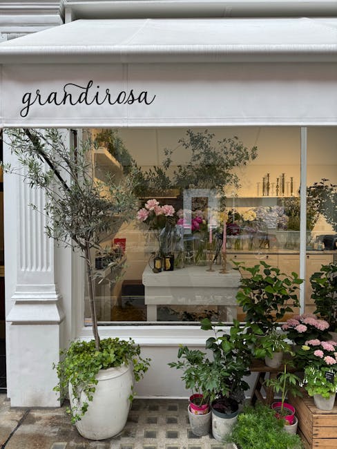 A flower shop exterior with a white awning displaying the word 'grandiroza' in black cursive lettering. In front of the shop, there are potted plants including an olive tree in a large white pot, pink