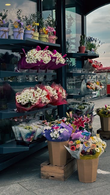 A display of various floral arrangements and bouquets at a flower shop, featuring fresh white daisies, pink roses, purple lilies, and mixed seasonal flowers. The flowers are wrapped in colorful tissue