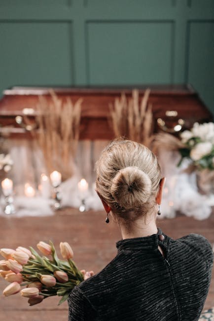 A woman with platinum blonde hair styled in a neat bun, wearing a black textured outfit, is holding a bouquet of soft pink tulips with green stems and leaves. She stands indoors in front of a rustic w