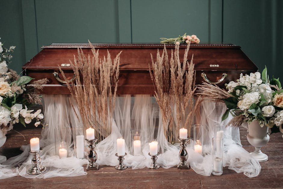 A floral arrangement set against a dark green wall, featuring a series of candles in silver holders, white fairy lights, and tall dried grasses in front of a closed wooden casket. Flanking the casket 