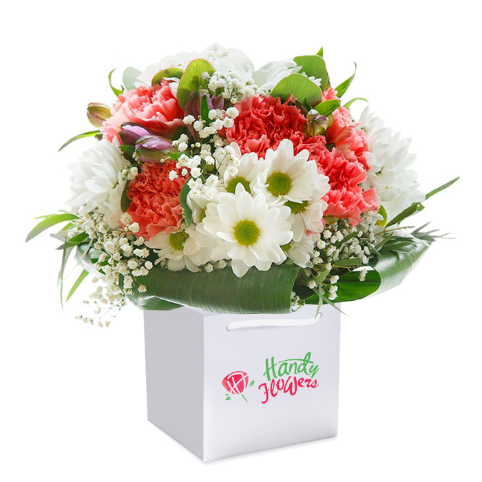 Captured in meticulous detail, this luxurious floral arrangement stands upright within a flawless, square white gift bag-its elegance heightened by the clean, minimalist backdrop that enhances the bursting vibrancy within. The bouquet boldly combines soft coral and blush-pink carnations, the ruffled symmetry of their petals lending a sumptuous, tactile depth that feels as inviting as a summer walk along Hampton's tranquil streets. Between the carnations, clusters of pure white daisies beam gently, their golden-green eyes creating a cheerful focal point that recalls the lively community spirit seen on Hampton High Street. Wisps of baby's breath interweave among the larger flowers, evoking clouds drifting across open skies above Hampton Court. Subtle unopened buds tinged with purple suggest new beginnings and fresh surprises, peeking from the mosaic of glossy, deep green leaves that encircle the base in a protective embrace reminiscent of Hampton's verdant gardens. The robust white ribbons arching from the bag's top ensure secure handling for direct, thoughtful delivery. This exquisitely arranged bouquet, imbued with a sense of celebration and gentle sophistication, is ideal for marking birthdays, anniversaries, or spontaneous acts of kindness. For anyone in Hampton searching for a memorable floral gift that radiates freshness and local character, this image truly captures the essence of dedicated, community-rooted floristry.